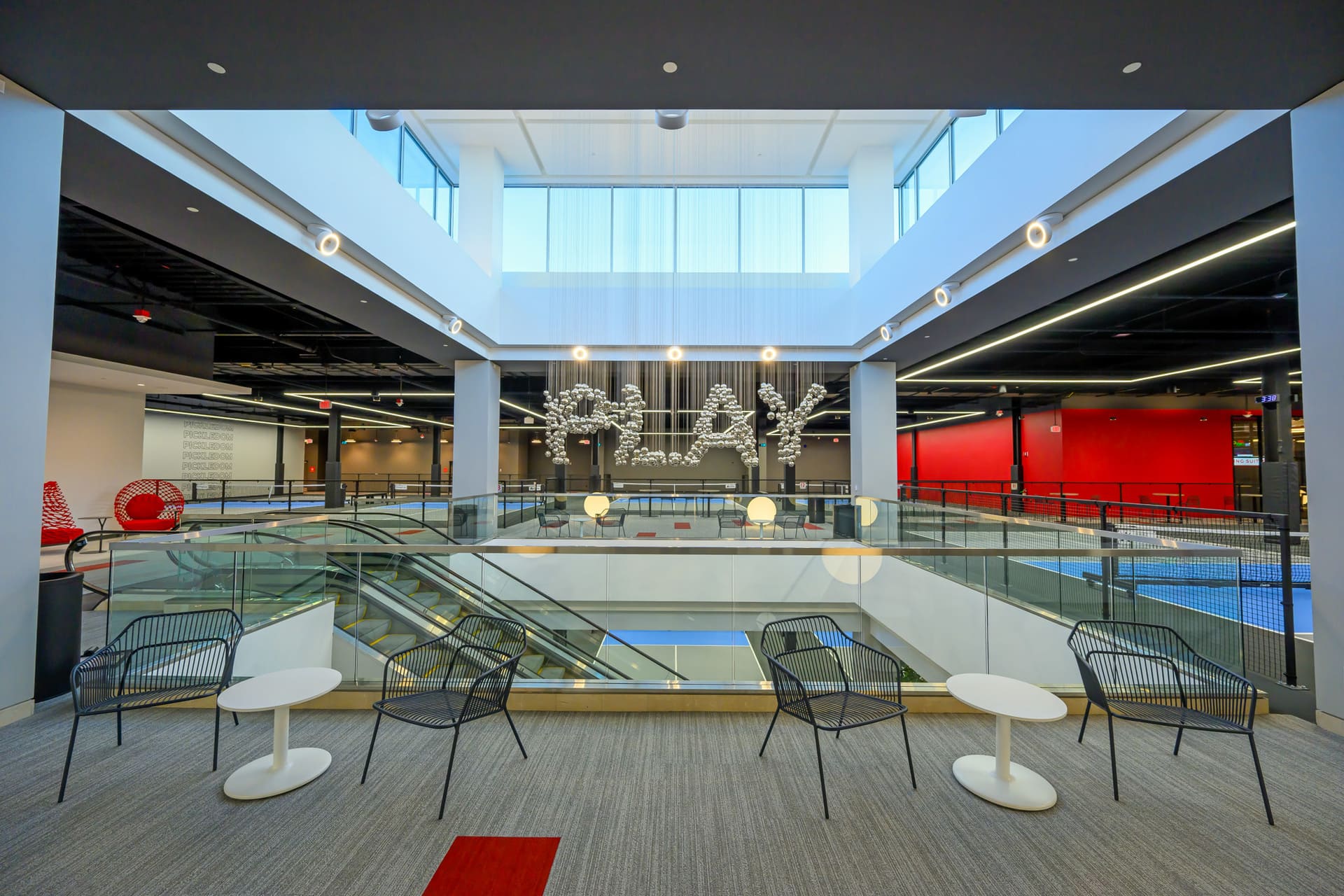 An open and bright atrium area with modern seating arrangements, a glass railing overlooking escalators, and a large decorative installation spelling "PLAY" made of spherical elements, surrounded by red and black accents.