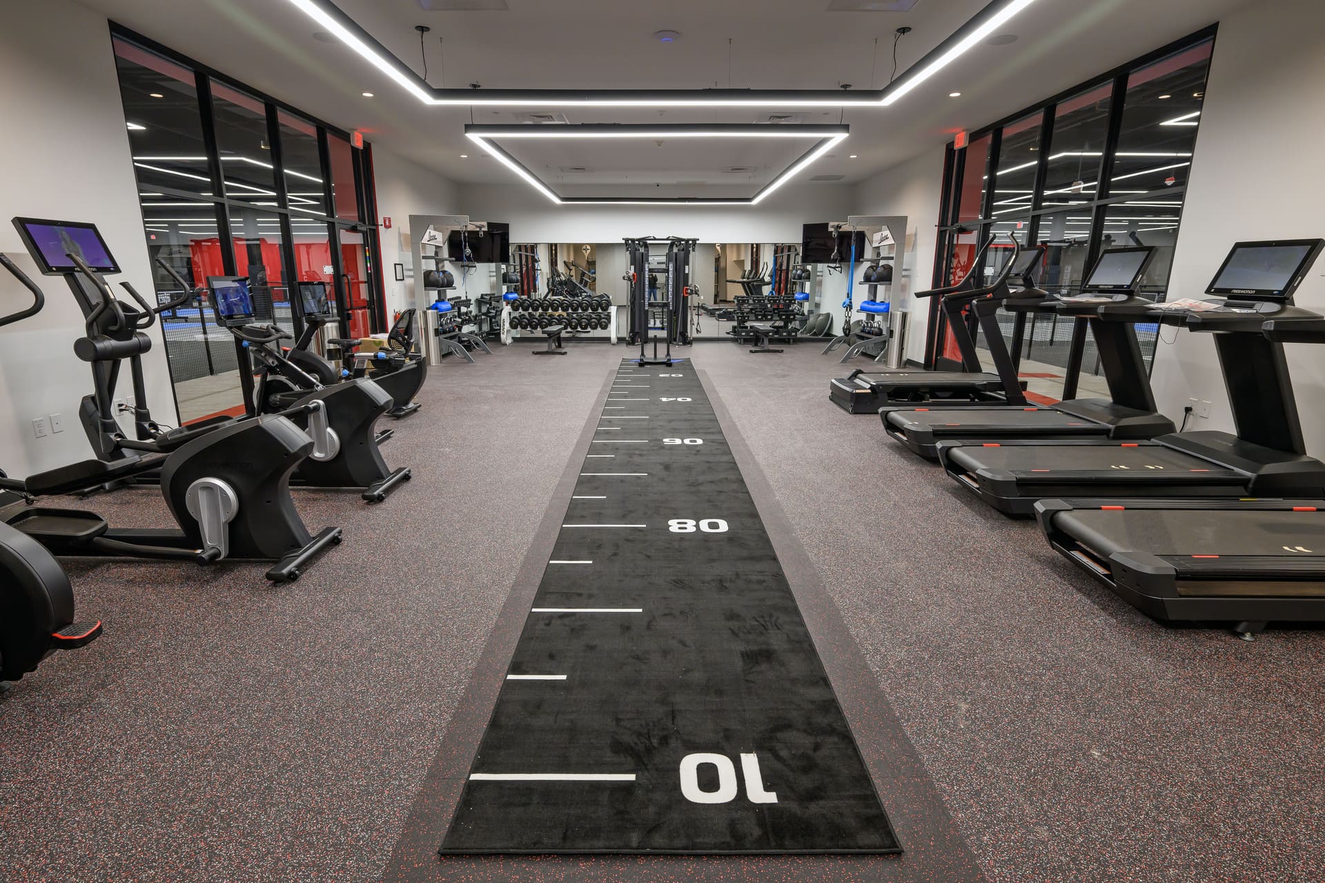 A modern fitness center featuring treadmills, elliptical machines, weight racks, and a long black agility mat