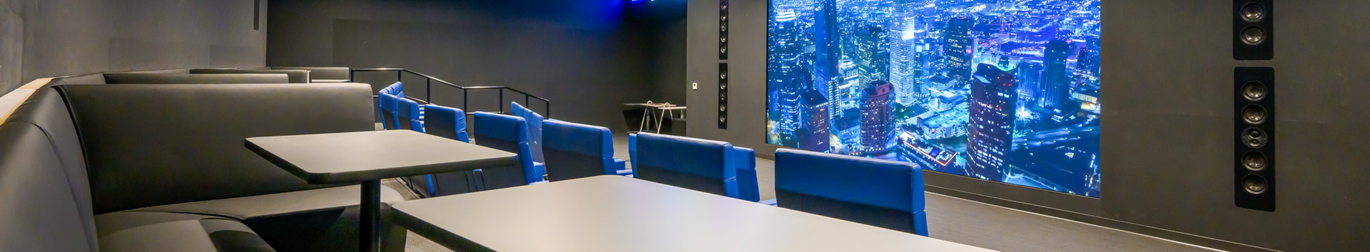 Invictus sports theatre features rows of blue seats, black booth-style seating, tables, and a large screen. The space includes wall-mounted speakers for an immersive viewing experience.