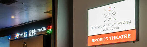 A modern sports theatre featuring a large screen displaying a cityscape, digital sports scores along the top, and a sign for "Invictus Technology Solutions Sports Theatre." The sleek space includes a minimalist design with dark walls and clean furnishings.