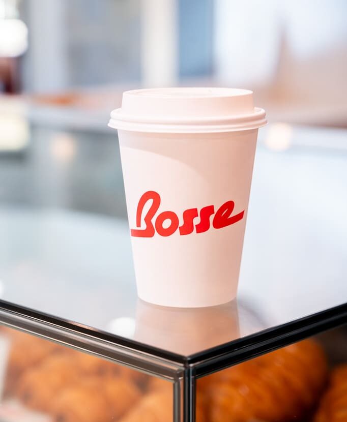Hot coffee in a Bosse Café cup placed on a glass display case with croissants underneath