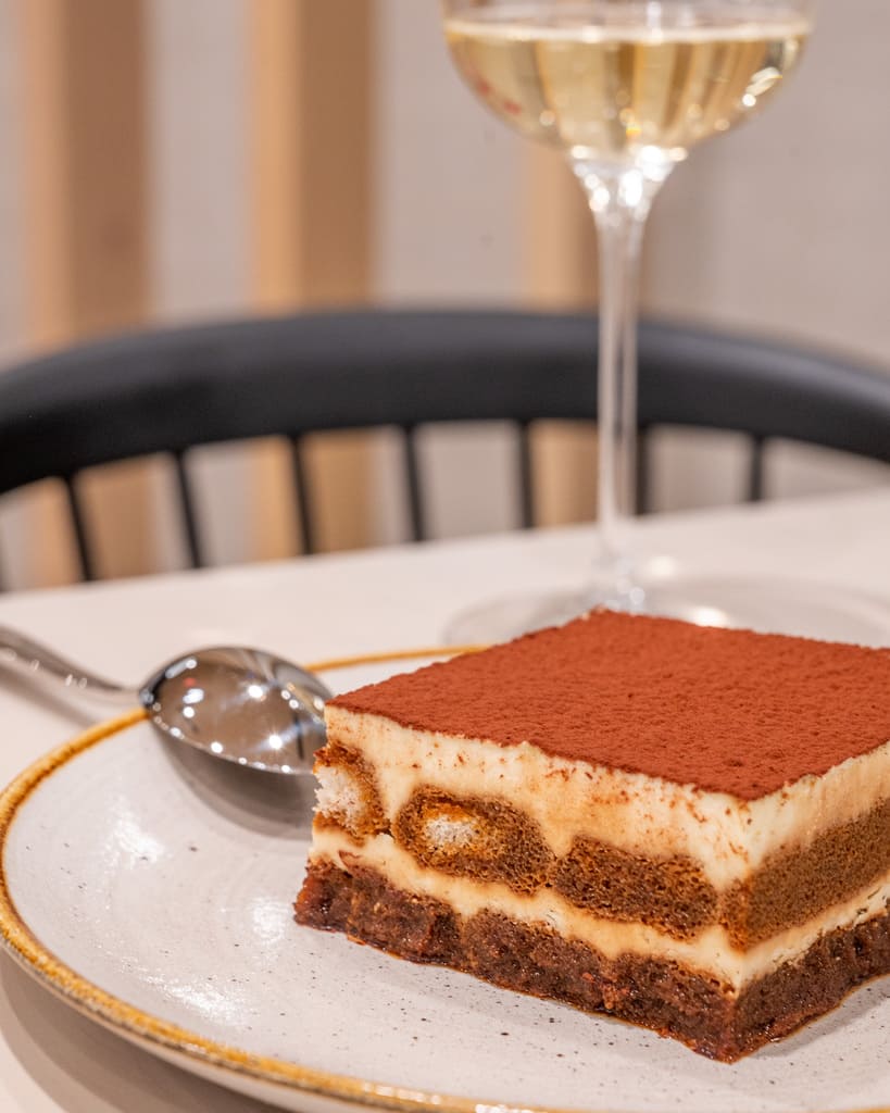 Classic tiramisu slice on a white plate beside a dessert spoon and glass of white wine