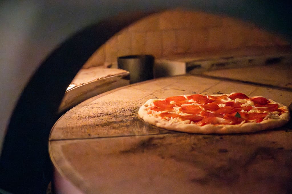 Pepperoni pizza baking inside a stone wood-fired oven, with bubbling cheese and crispy edges forming under the oven’s heat.