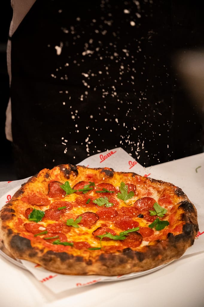 Freshly baked pepperoni pizza with charred crust and fresh herbs on a branded sheet of paper, set on a white surface