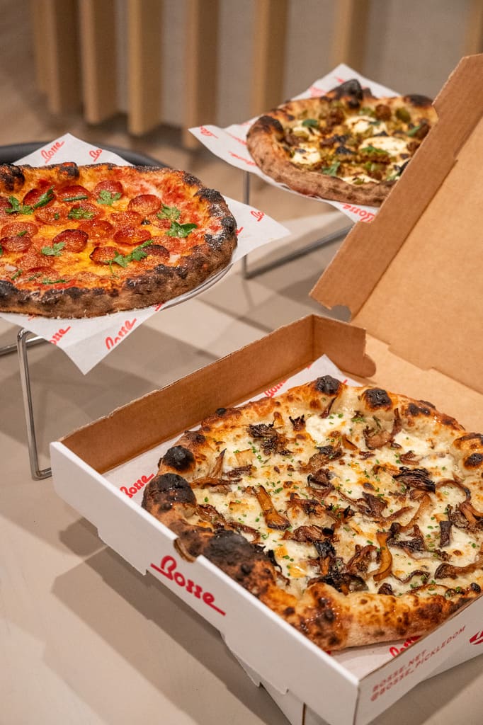 Three artisan-style pizzas displayed on stands and in a box, featuring pepperoni, white mushroom, and roasted vegetable toppings with charred crusts.