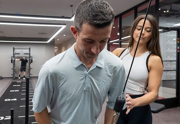 Male client performs cable exercises under supervision from a female personal trainer in a gym.