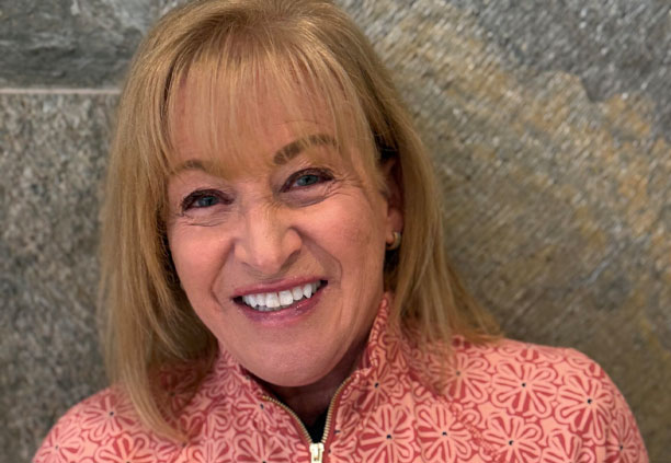 Smiling woman with blond hair wearing a patterned pink zip-up jacket, posing in front of a textured wall.