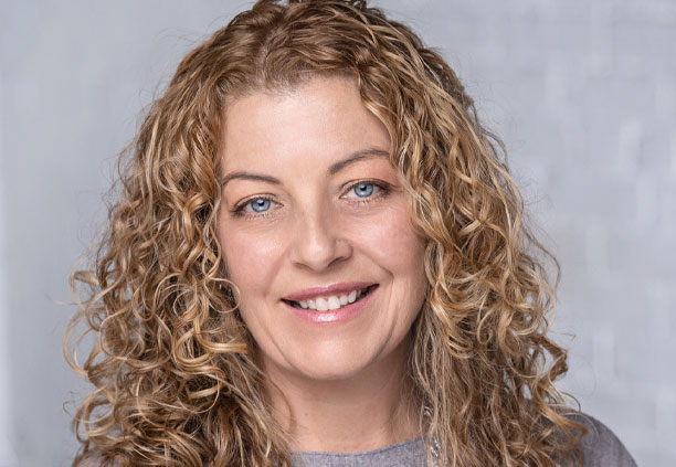 Woman with curly blonde hair and blue eyes smiles in a professional portrait with a light gray background.